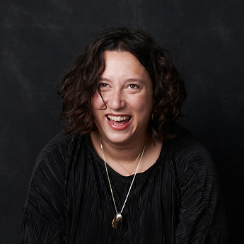 A headshot photo of Cayn Rosmarin. Cayn has brown short curly hair. Cayn is pictured smiling in front of a black background and is wearing a black shirt with a silver necklace.