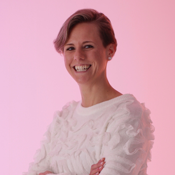 A headshot of Giulia Giorgi. Giulia Giorgi has blonde short hair and is smiling. She is standing against a pink background and is wearing a white long sleeve shirt and has her arms crossed.