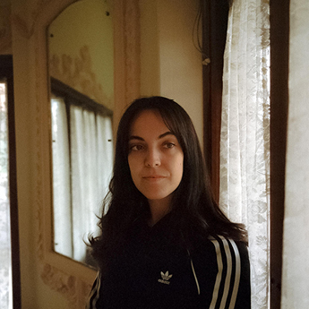 A woman with brown hair stands in the light of a window wearing a black tracksuit jumper.
