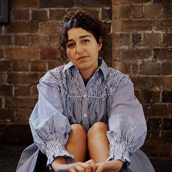 A woman with brown hair sits facing the camera with her arms clasped around her knees. She is wearing a blue and white striped dress.