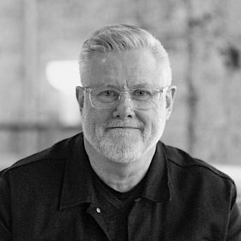 A black and white headshot image of Simon Spellicy. Simon has grey hair and is wearing glasses. He is wearing a dark collared shirt.