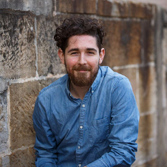 A headshot image of Hayden Walsh. Hayden has a blue business shirt on and has short dark curly hair. He has a beard and is smiling at the camera while leaning against a brick wall.